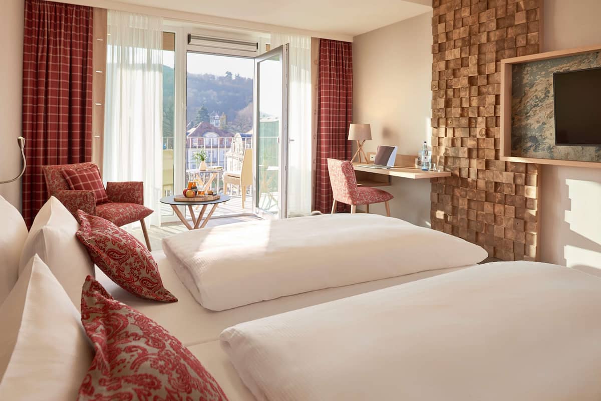 Une chambre d’hôtel avec deux lits, un bureau avec un ordinateur portable, une chaise, une petite table avec une plante et une vue sur l’extérieur.