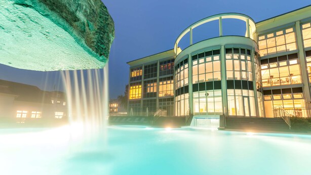 A modern building with a circular glass structure, a pool, and a waterfall feature.
