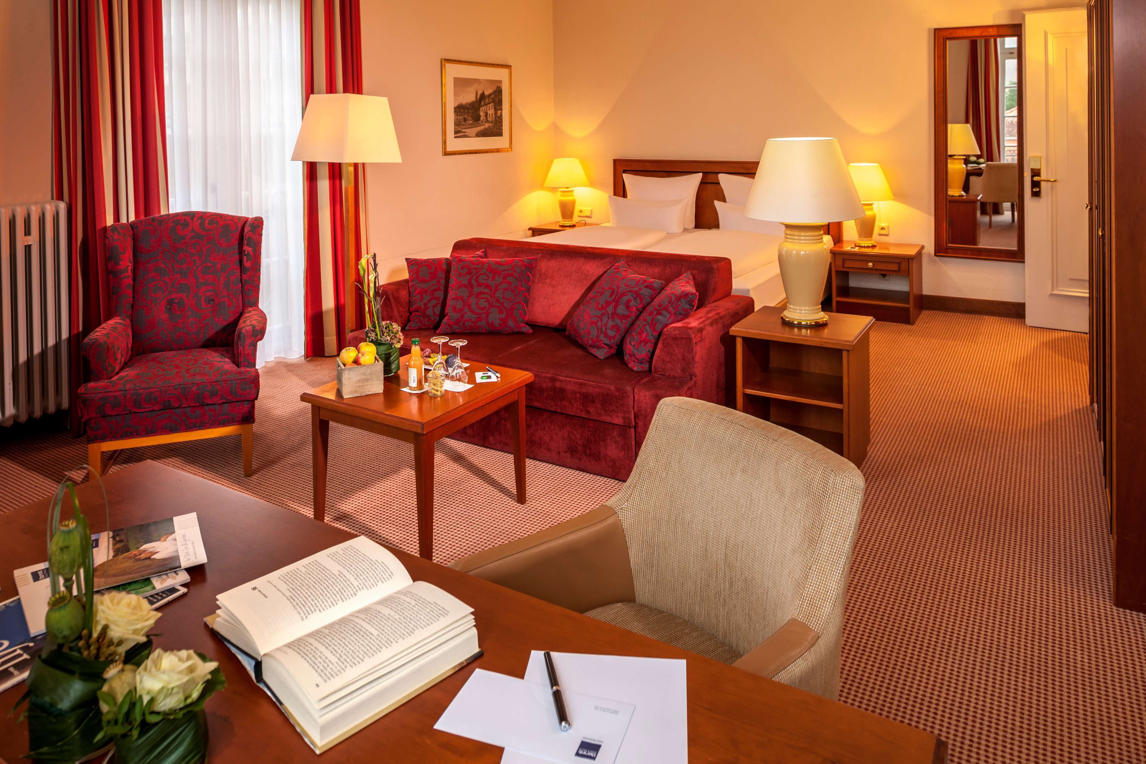 A hotel room with a red patterned armchair, a red sofa, a wooden desk with a book and papers, a small table with a vase of flowers, and a bed with white linens.