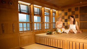 A sauna room with wooden benches, large windows, and a small table with fruits and a drink.