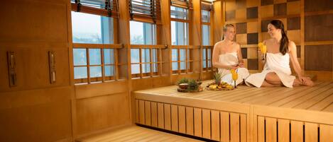 A sauna room with wooden benches, large windows, and a small table with fruits and a drink.