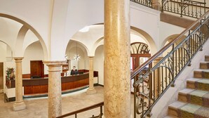 A lobby with marble columns, a reception desk, and a staircase with ornate metal railings.