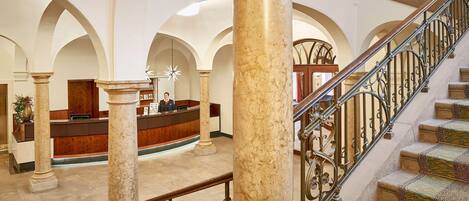 A lobby with marble columns, a reception desk, and a staircase with ornate metal railings.