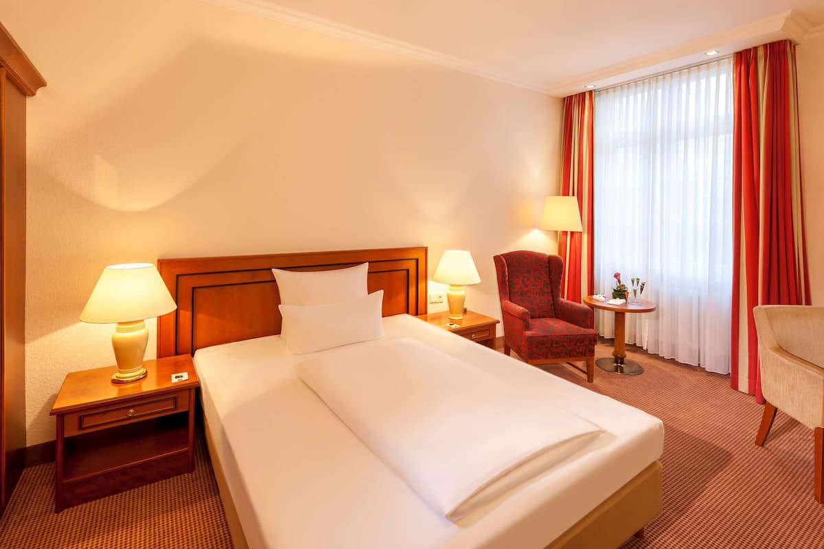 Une chambre d’hôtel avec un grand lit, deux tables de chevet avec des lampes, une chaise, un bureau et une fenêtre avec des rideaux.