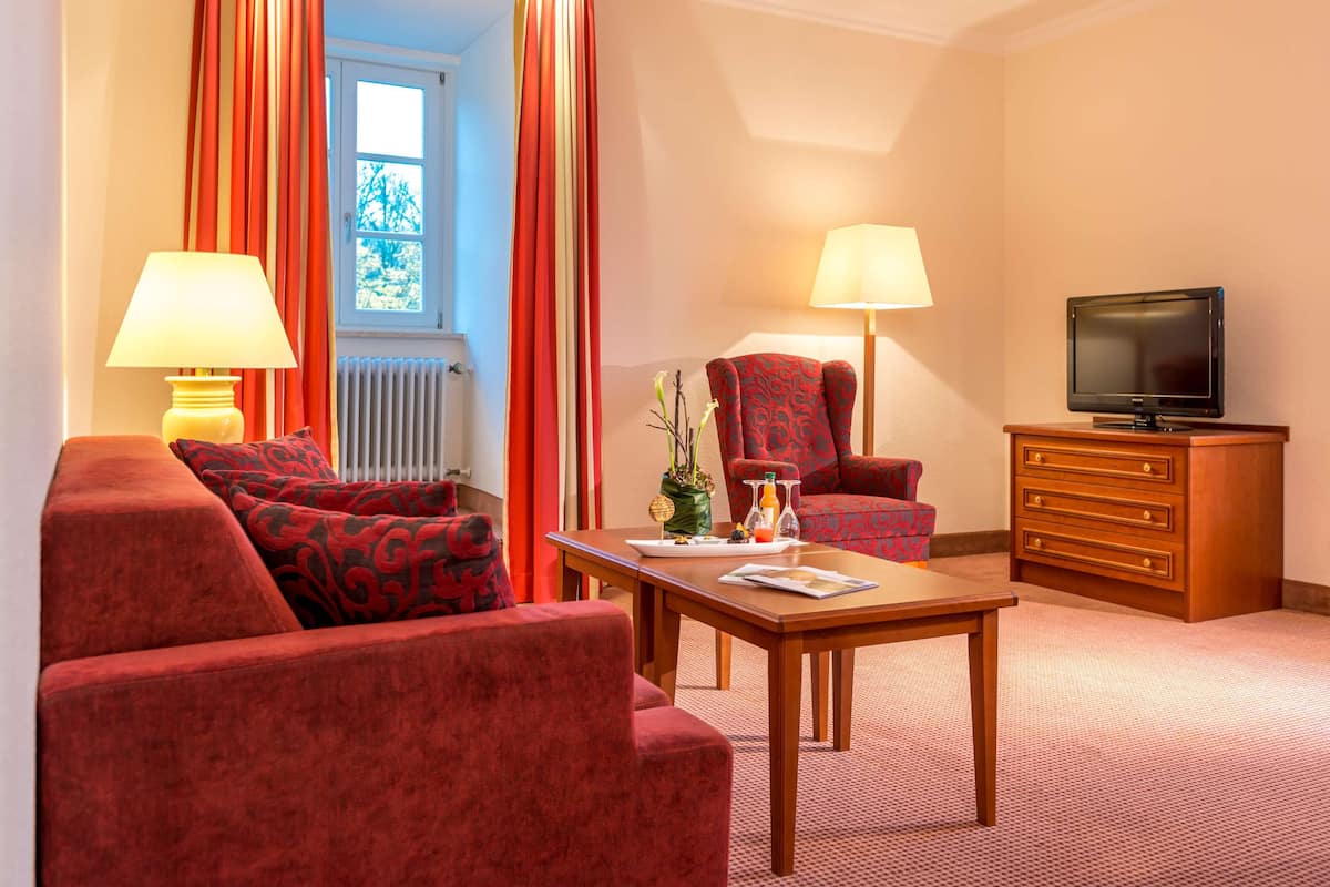Une chambre d’hôtel avec un canapé rouge recouvert de tissu, un fauteuil assorti, une table basse en bois, un téléviseur posé sur un meuble en bois et deux lampes.