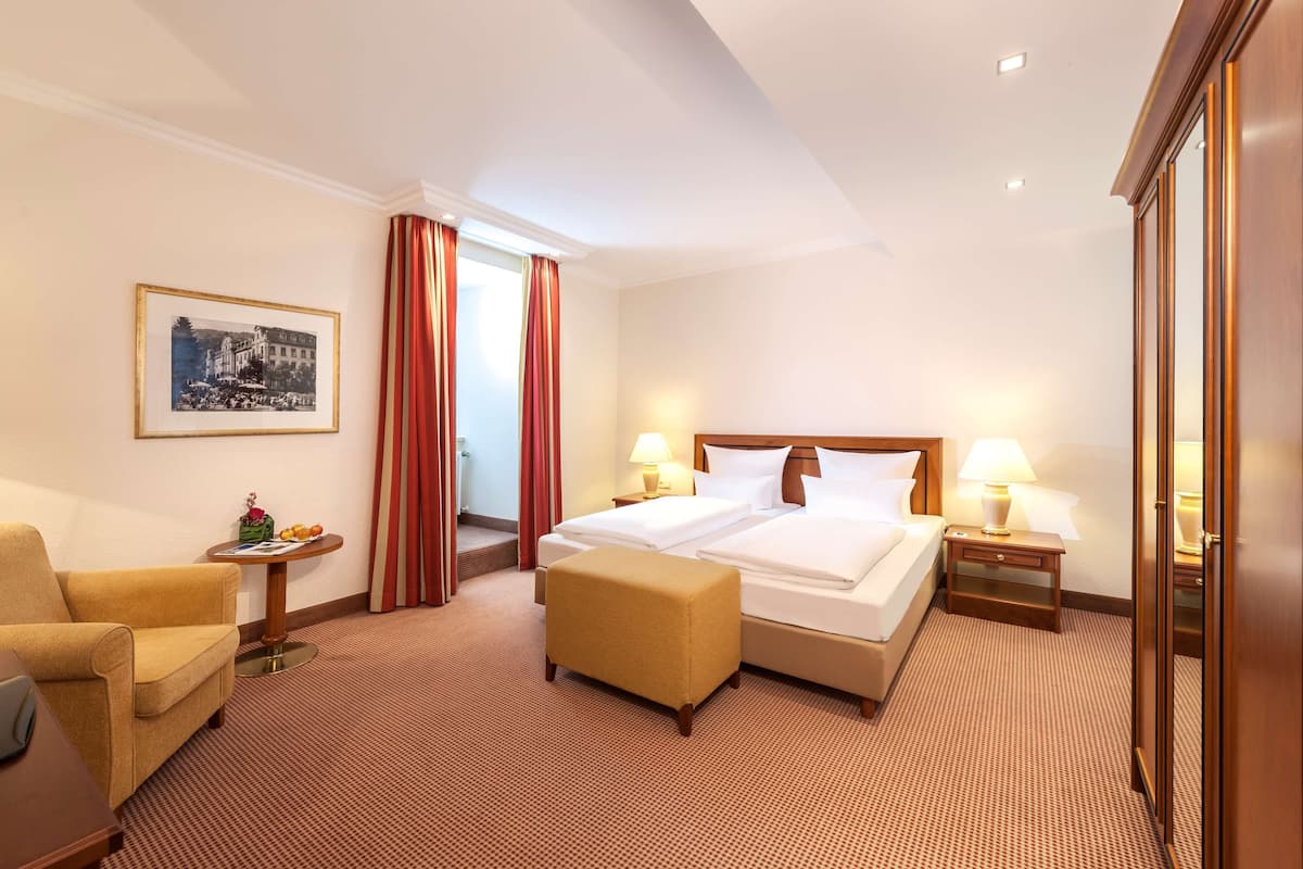 Une chambre d’hôtel avec un lit, une chaise, une petite table avec une lampe, un tableau au mur et une porte.