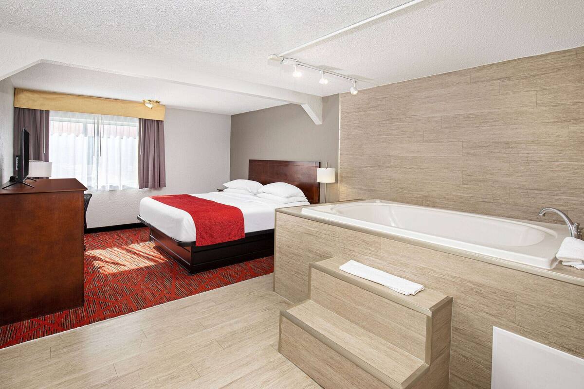 Ett hotellrum med en säng, ett skrivbord, ett stort fönster med gardiner, ett badrum med badkar och en röd matta.