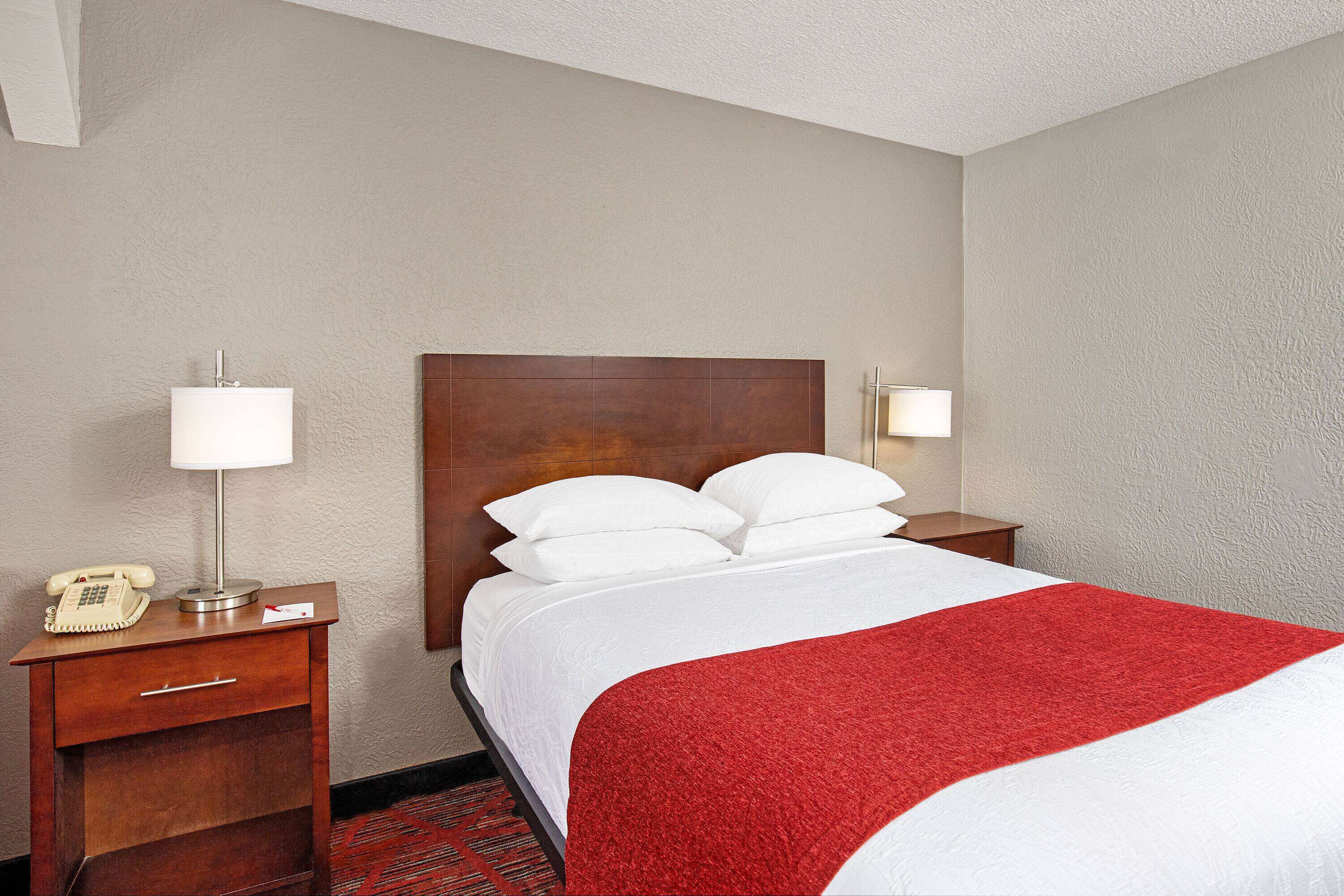 A hotel room with a neatly made bed, a wooden nightstand with a lamp, a telephone, and a red and white bedspread.