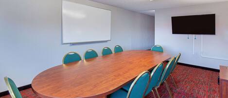 Uma sala de conferências com mesa de madeira, cadeiras azuis e um quadro branco, além de uma TV de tela plana.