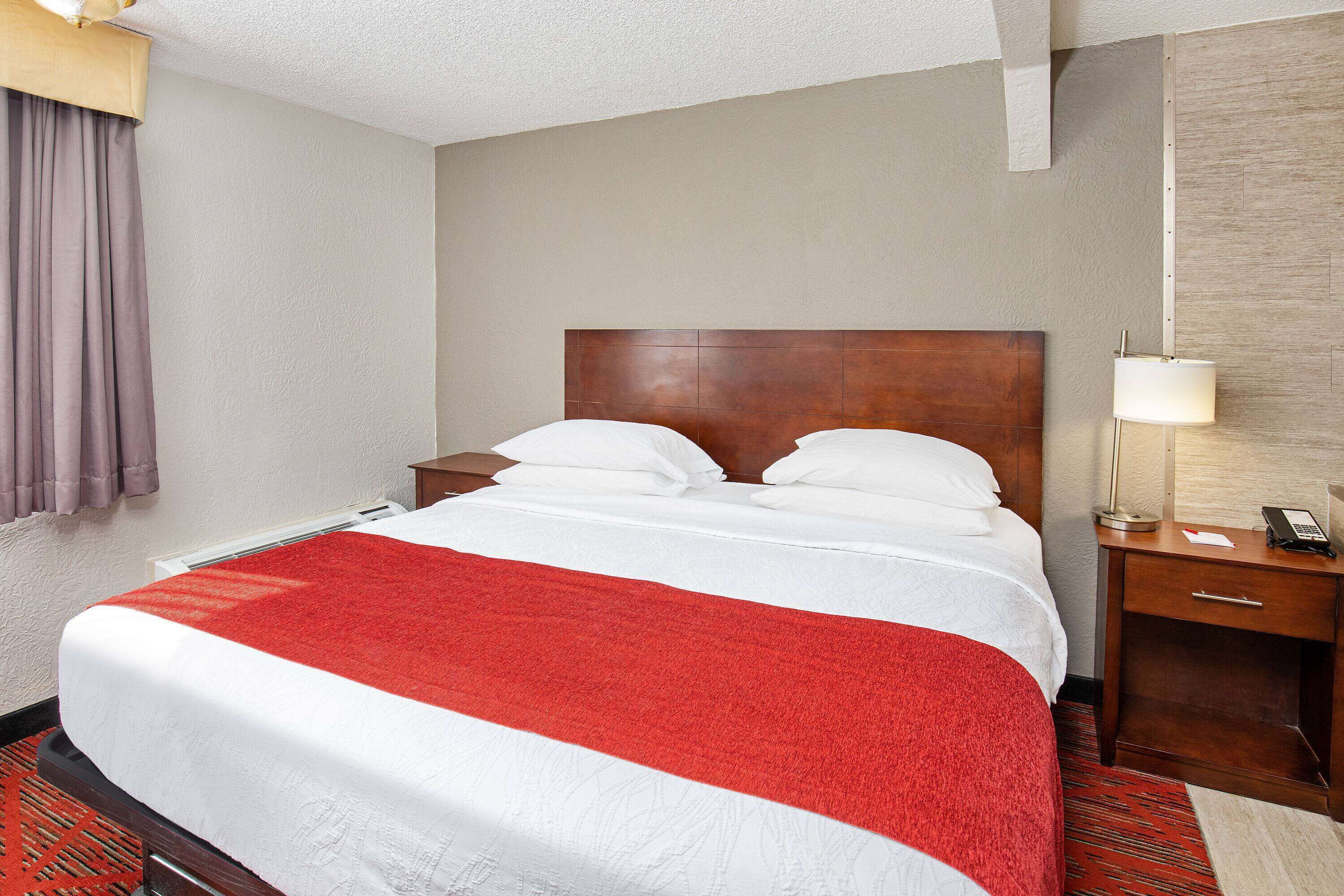 Een hotelkamer met een groot bed, een houten hoofdbord, een nachtkastje met een lamp en een rood-witte sprei.