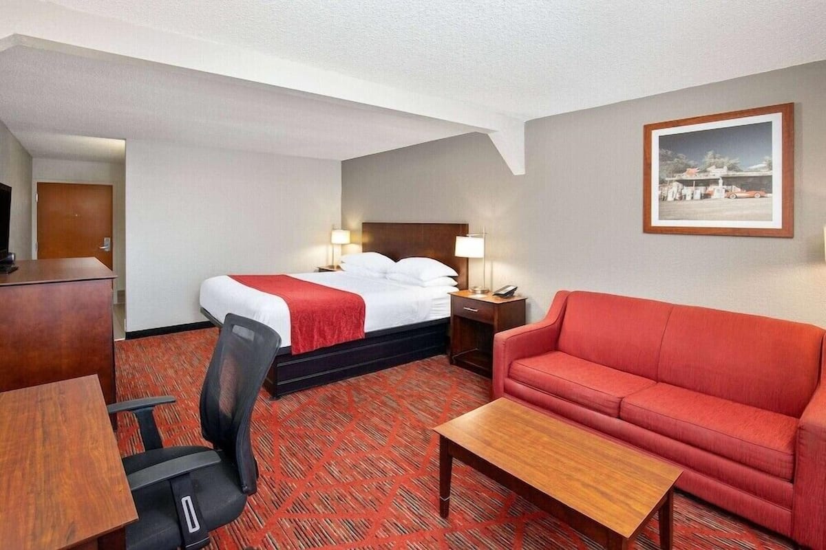 A hotel room with a bed, a red sofa, a wooden table, a desk, and a chair.
