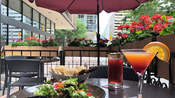 Mesa dispuesta con ensalada, un martini y pan, bajo un paraguas rojo, con vista a la ciudad de fondo.