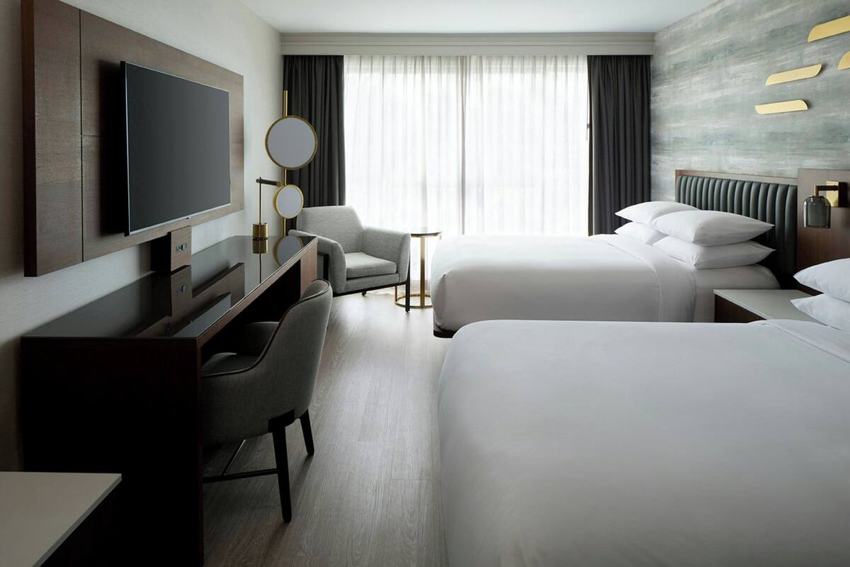 A hotel room with a TV, two beds, a desk, a chair, a small table, and a window with curtains.