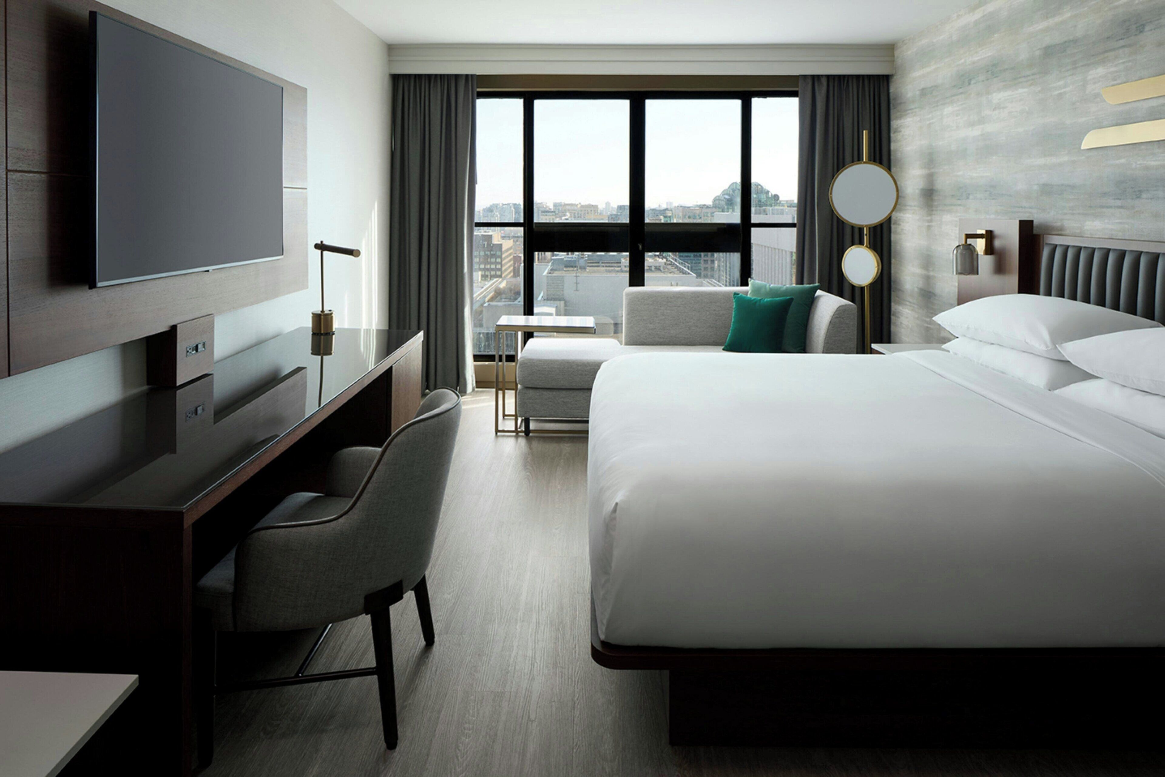 A hotel room with a bed, a desk, a chair, a television, and a view of the city through large windows.