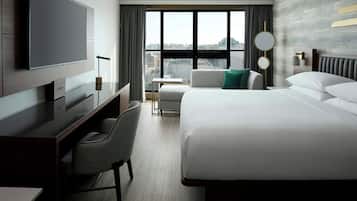 A hotel room with a bed, a desk, a chair, a television, and a view of the city through large windows.