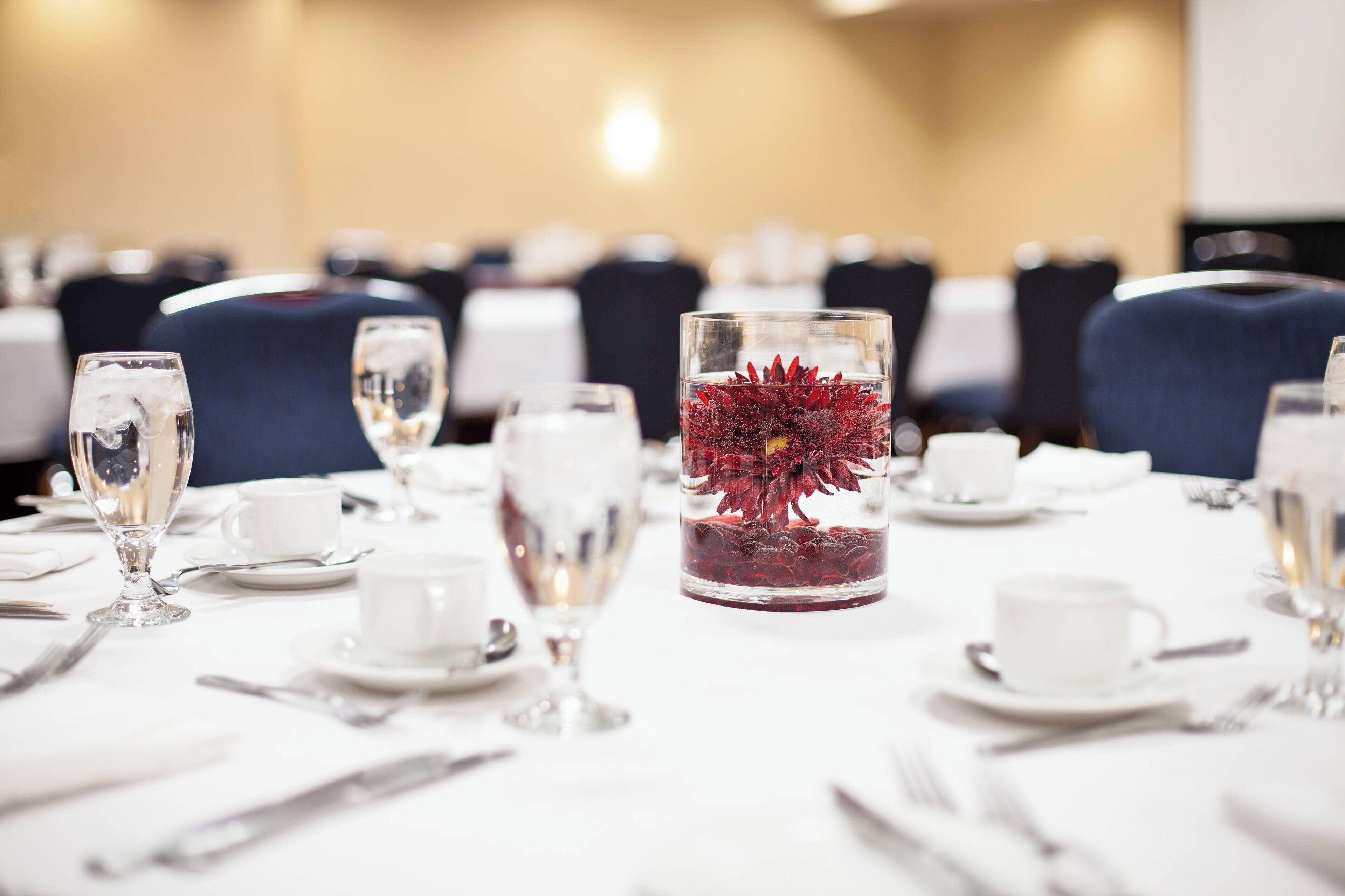 A dining table set with white tableware, including wine glasses, water glasses, and a centerpiece with red flowers in a glass container.