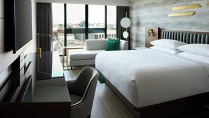 Habitación de hotel con cama, escritorio, silla, televisor, ventana con vista a la ciudad y espejo montado en la pared.