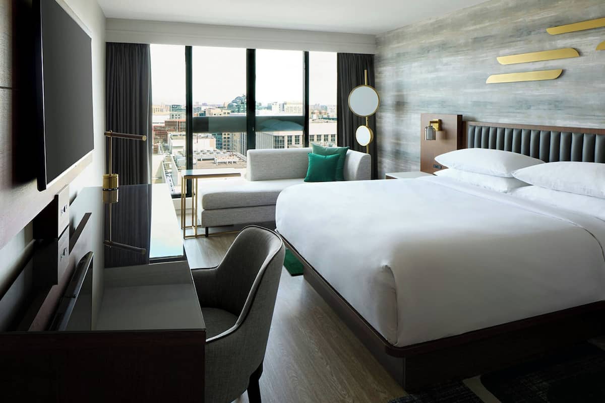 A hotel room with a bed, a desk, a chair, a TV, a window with a city view, and a wall-mounted mirror.