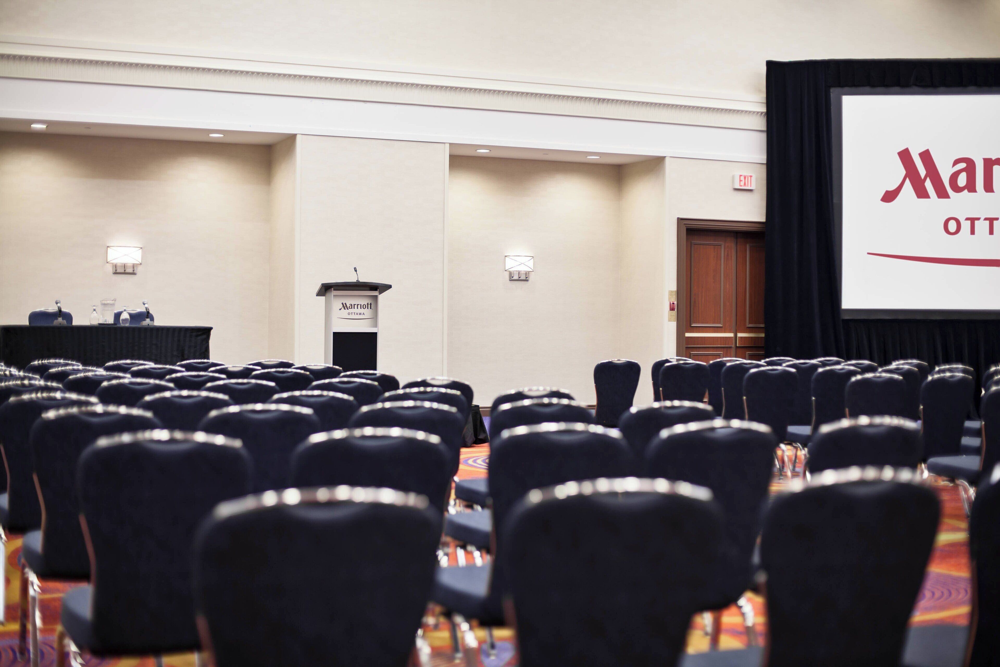 A conference room with rows of chairs, a podium, and a large screen displaying the Marriott logo.