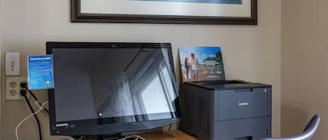 Un escritorio de madera con dos monitores, un teclado, un ratón e impresora. Encima del escritorio, un cuadro enmarcado.