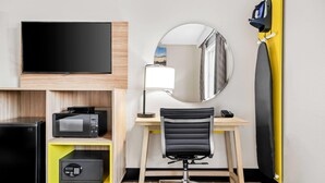 A hotel room with a flat-screen TV, a microwave, a safe, a desk with a chair, a mirror, and a yellow ironing board.
