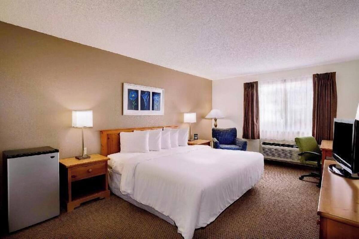 A hotel room with a bed, two bedside lamps, a desk with a television, a small refrigerator, and a window with curtains.