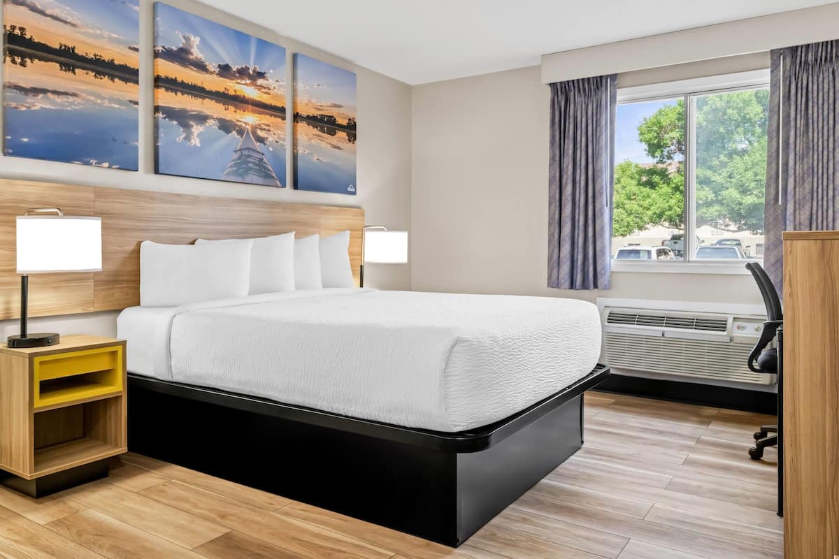 A modern hotel room with a large bed, two bedside lamps, a wooden nightstand, a view of the outdoors through a window, and a wall-mounted air conditioning unit.