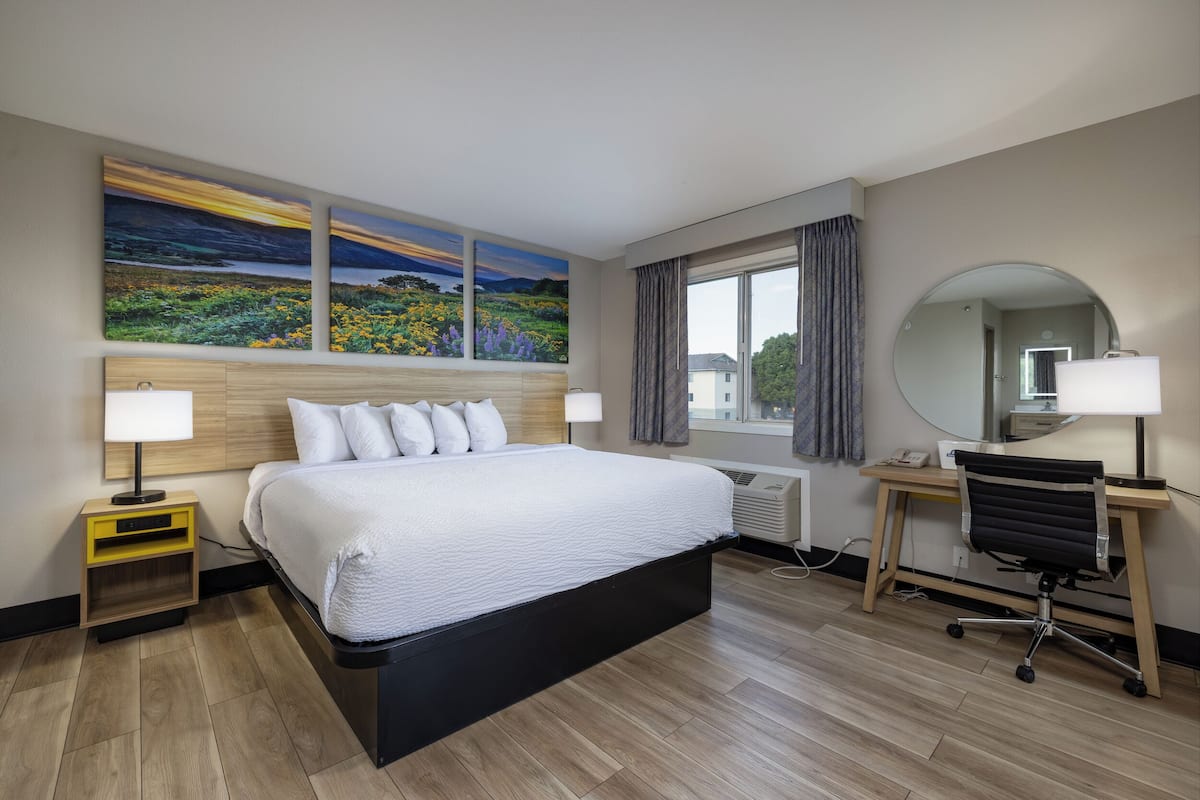A hotel room with a large bed, a desk with a chair, a round mirror, and a painting of a landscape on the wall.
