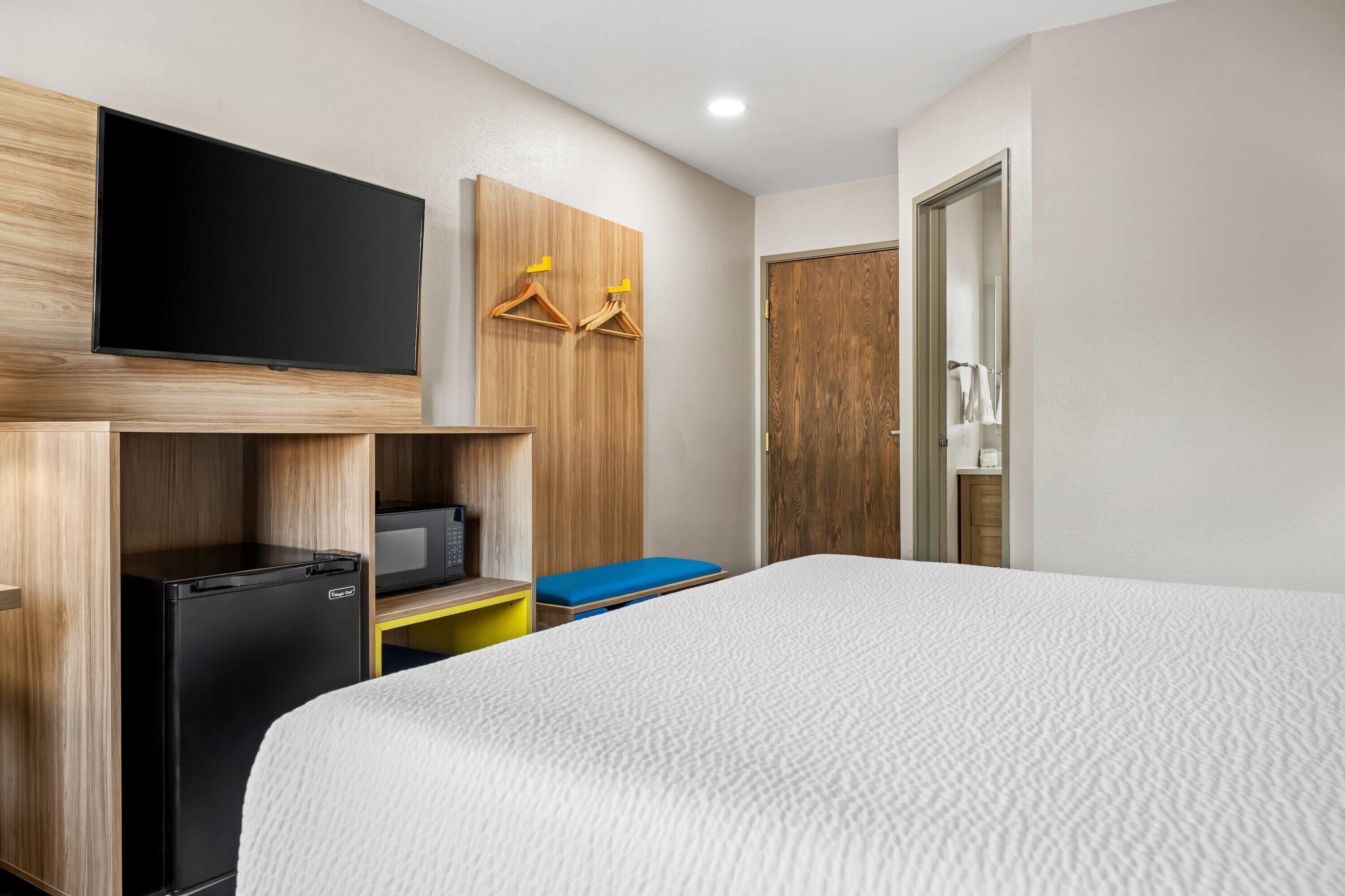 A hotel room with a bed, a television mounted on a wooden cabinet, a small refrigerator, and a wooden wardrobe with hangers.