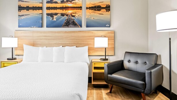 A hotel room with a large bed, two bedside tables with lamps, a black leather armchair, and three sunset paintings on the wall.