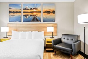A hotel room with a large bed, two bedside tables with lamps, a black leather armchair, and three sunset paintings on the wall. - Days Inn by Wyndham Billings (Billings)