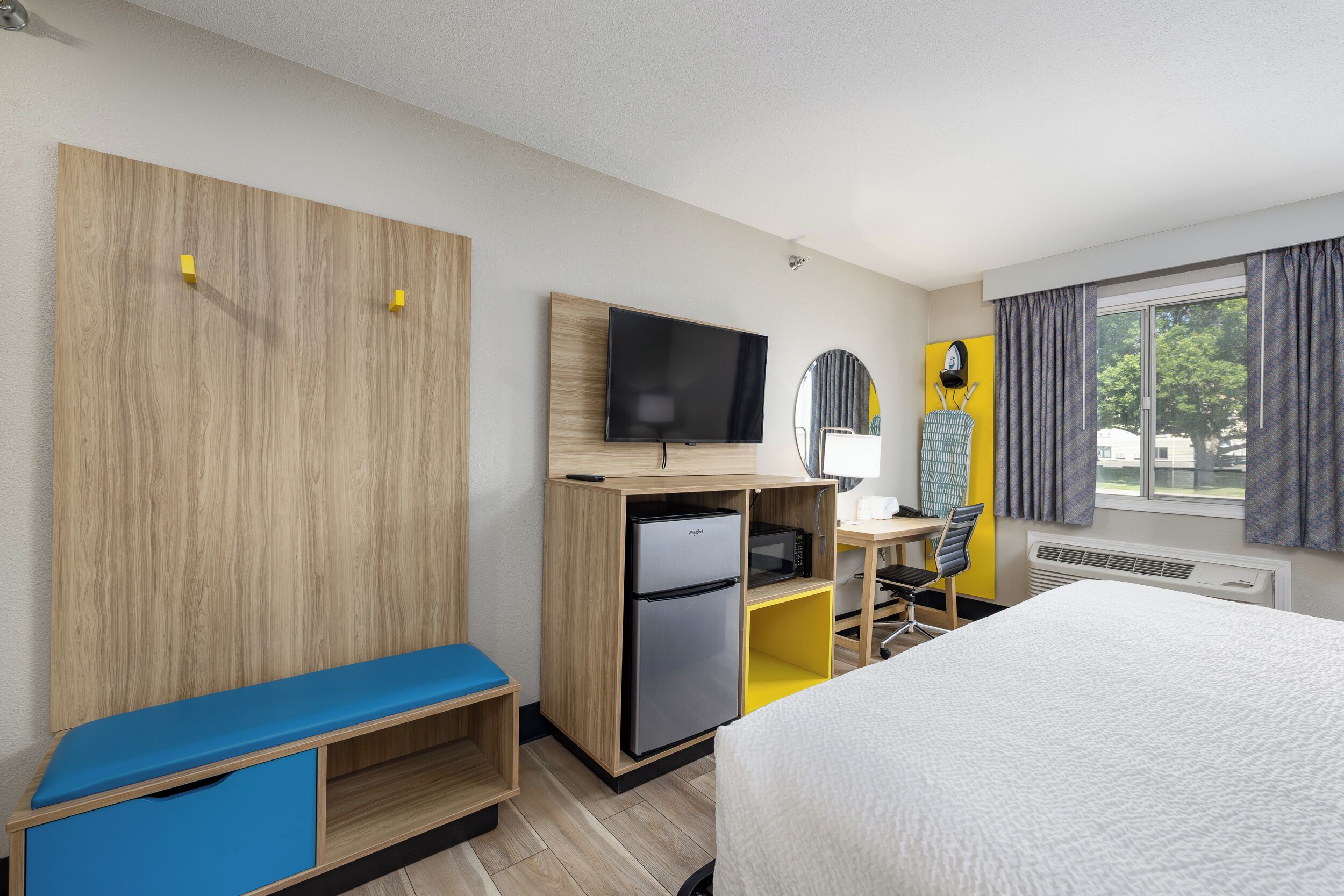 A hotel room with a bed, a desk with a computer, a TV, a mini-fridge, and a seating area.