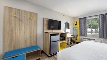 A hotel room with a bed, a desk with a computer, a TV, a mini-fridge, and a seating area.