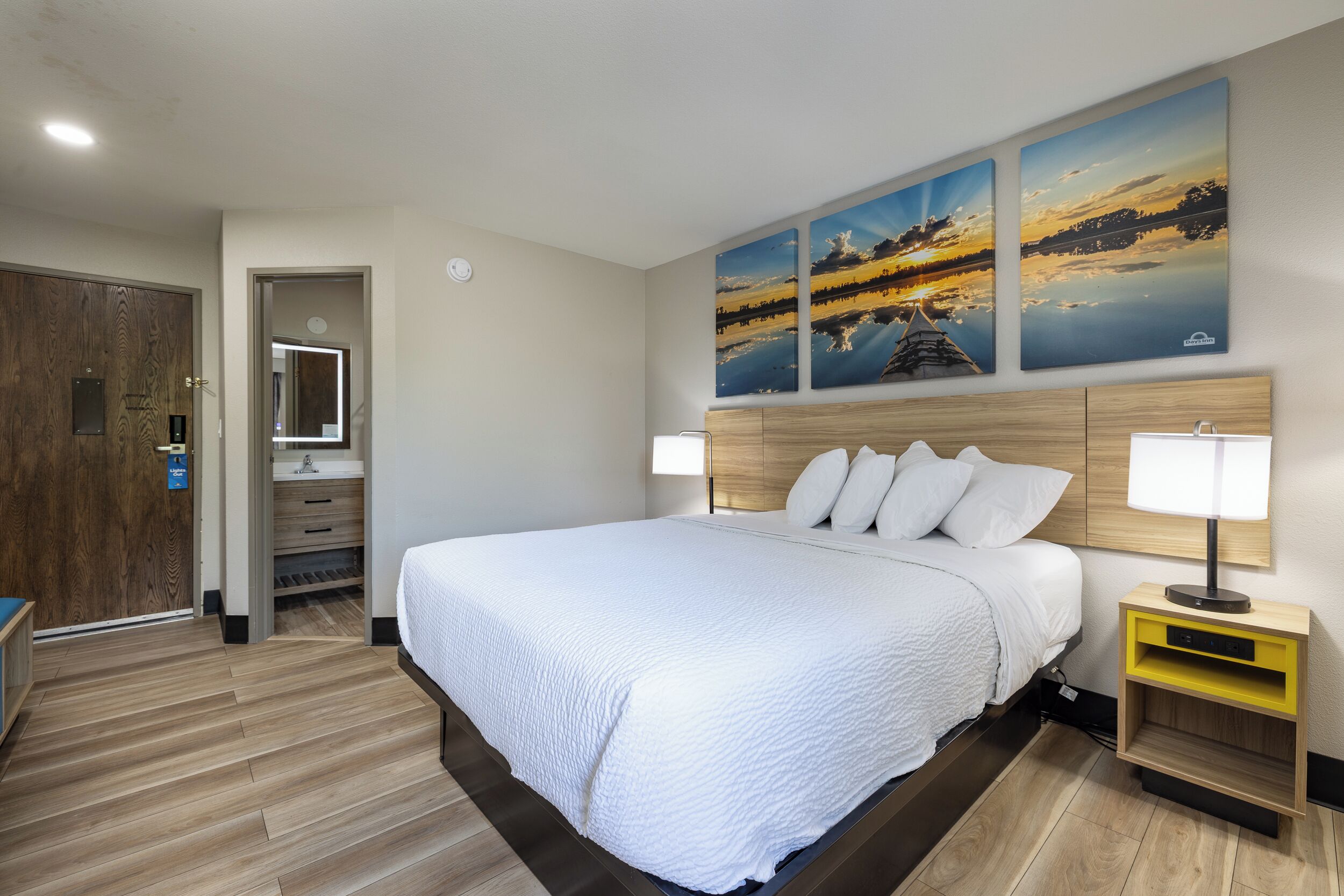 Een hotelkamer met een groot bed, houten vloer, een houten hoofdbord, een nachtkastje met een lamp en drie aan de muur hangende schilderijen van een zonsondergang.