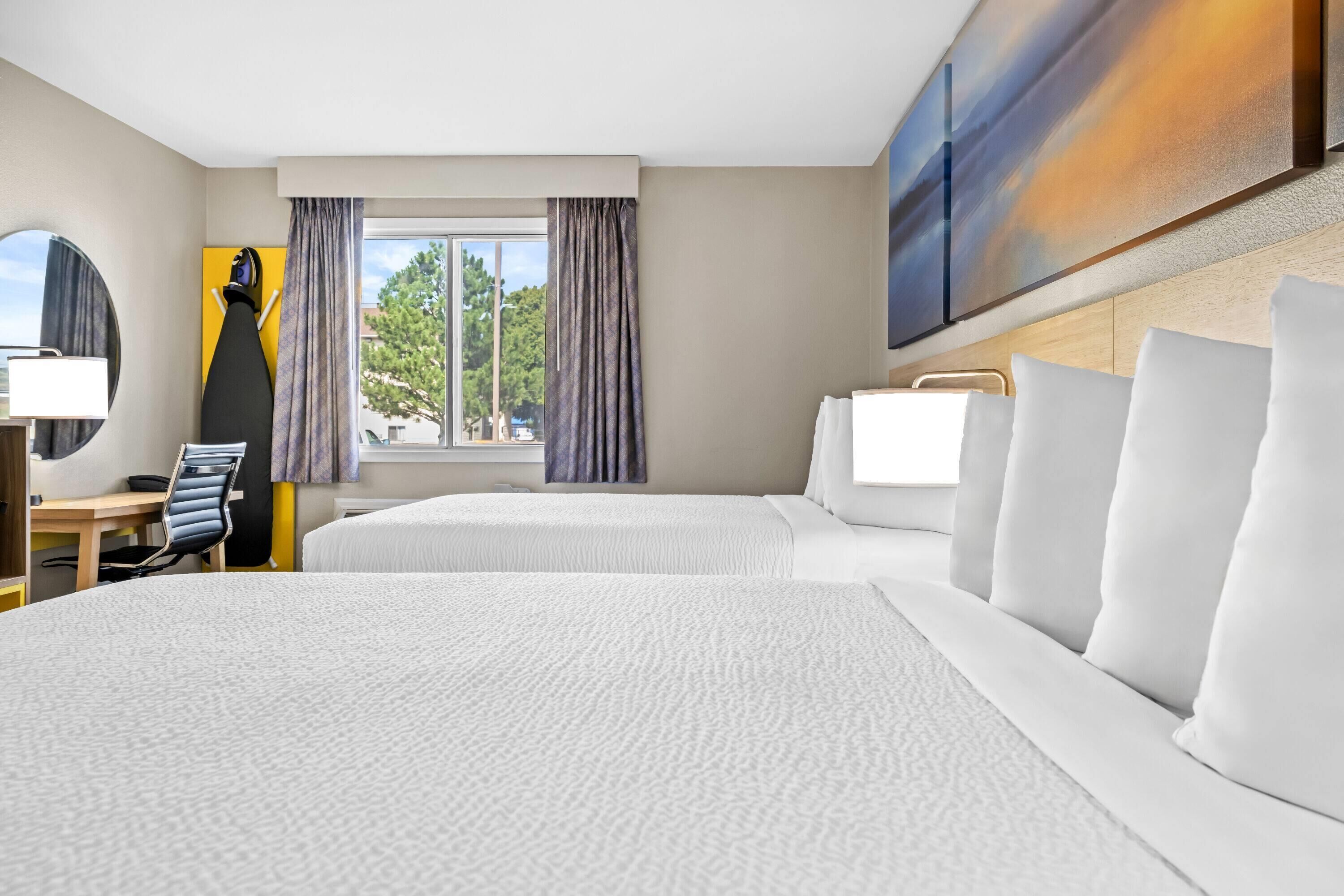 Habitación de hotel con dos camas, un escritorio, una silla, una lámpara y un ventanal grande con cortinas.
