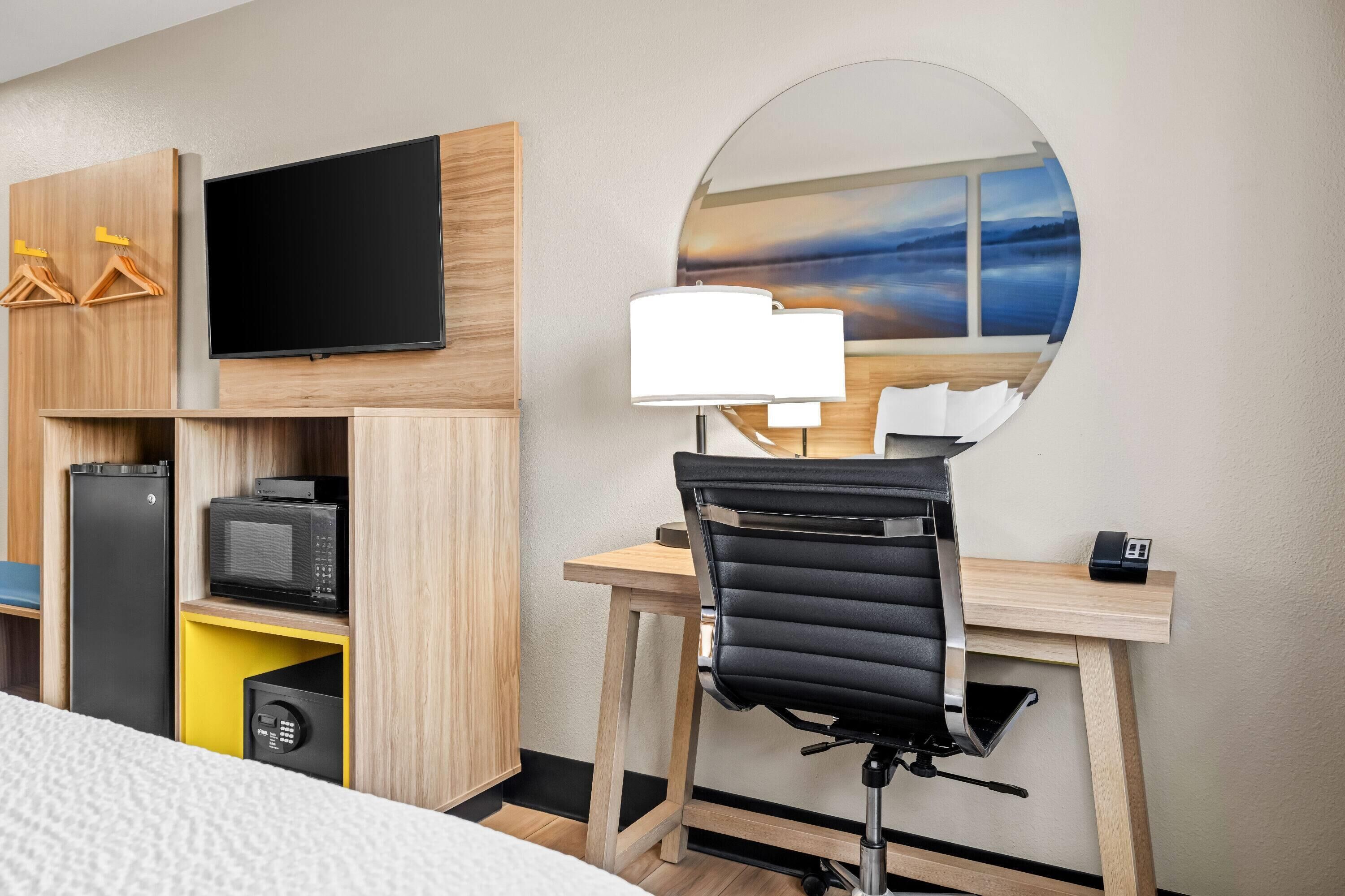 A hotel room with a bed, a desk with a chair, a flat-screen TV, a microwave, a refrigerator, and a large mirror.