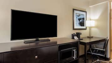 A hotel room with a large flat-screen TV, a desk with a chair, a framed picture on the wall, a lamp, and a coffee maker.