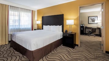 A hotel room with a large bed, two bedside tables with lamps, a desk with a chair, and a door leading to another room.