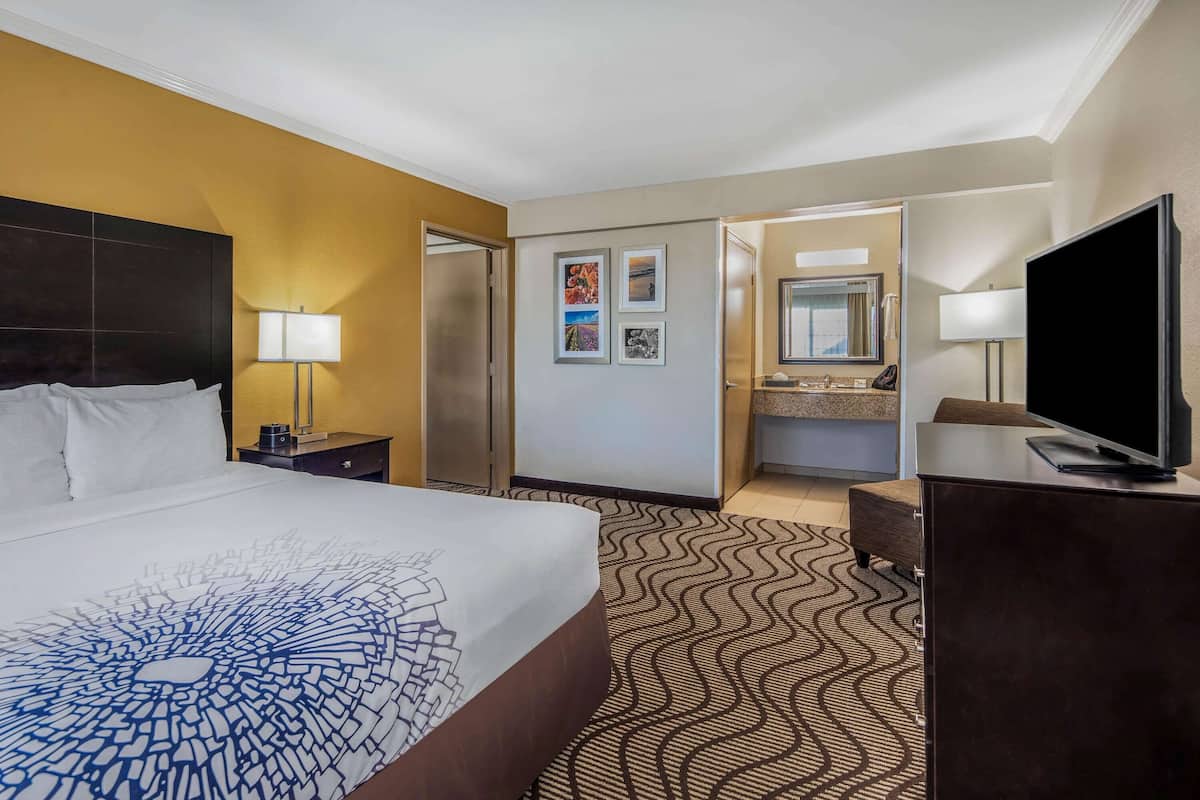 A hotel room with a large bed, a desk, a television, a bathroom with a sink and mirror, and a patterned carpet.