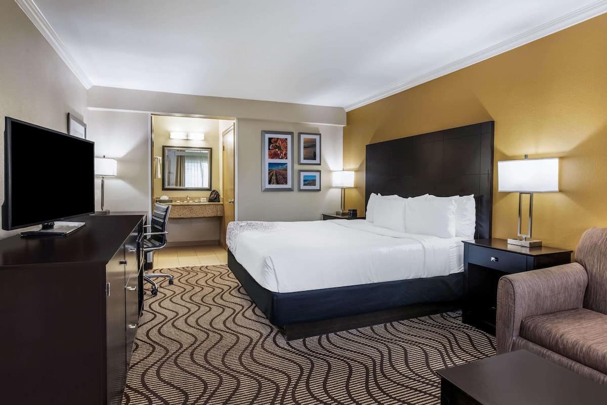 A hotel room with a large bed, a desk with a flat-screen TV, a chair, a nightstand with a lamp, and a sofa.