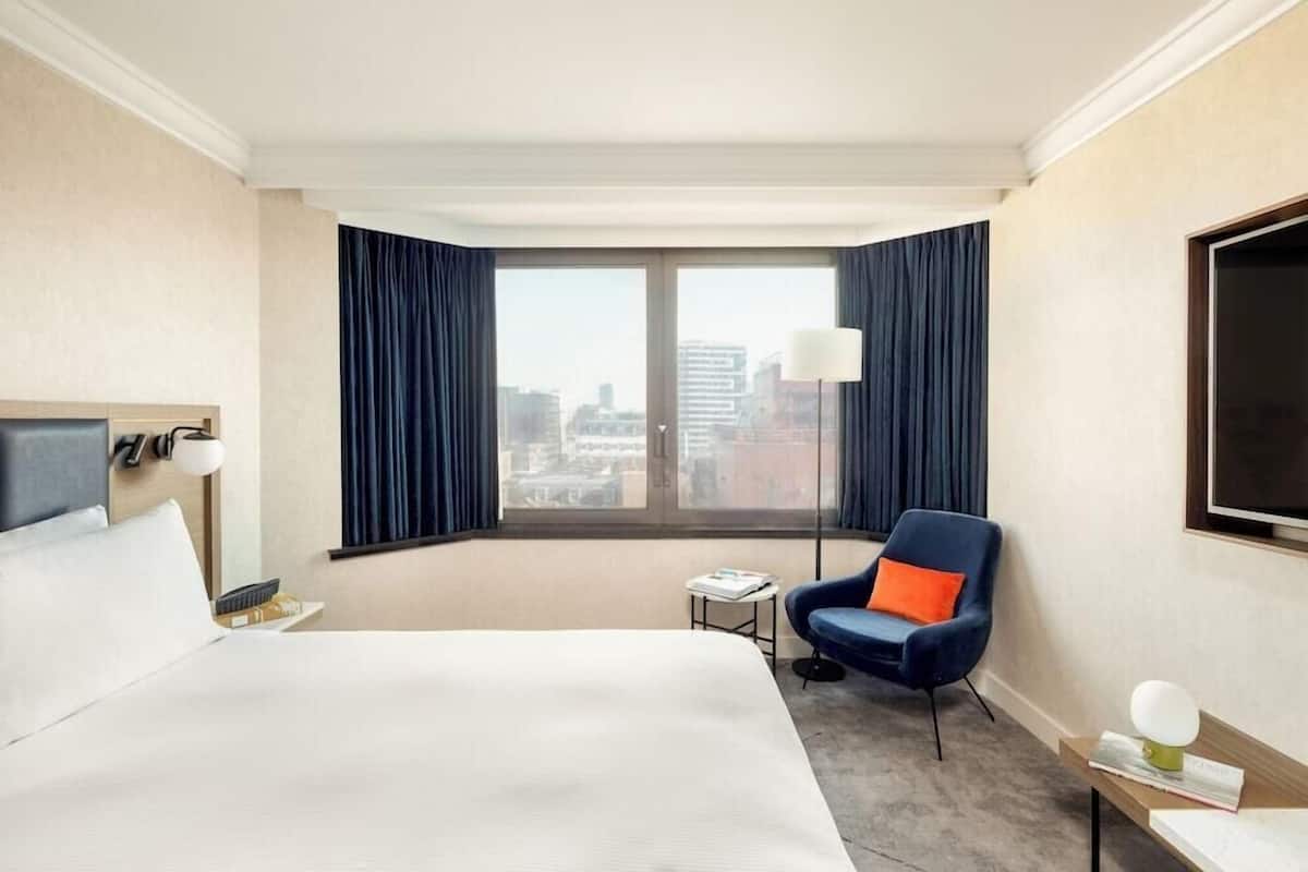 A hotel room with a bed, a chair, a small table, a lamp, and a large window with curtains.