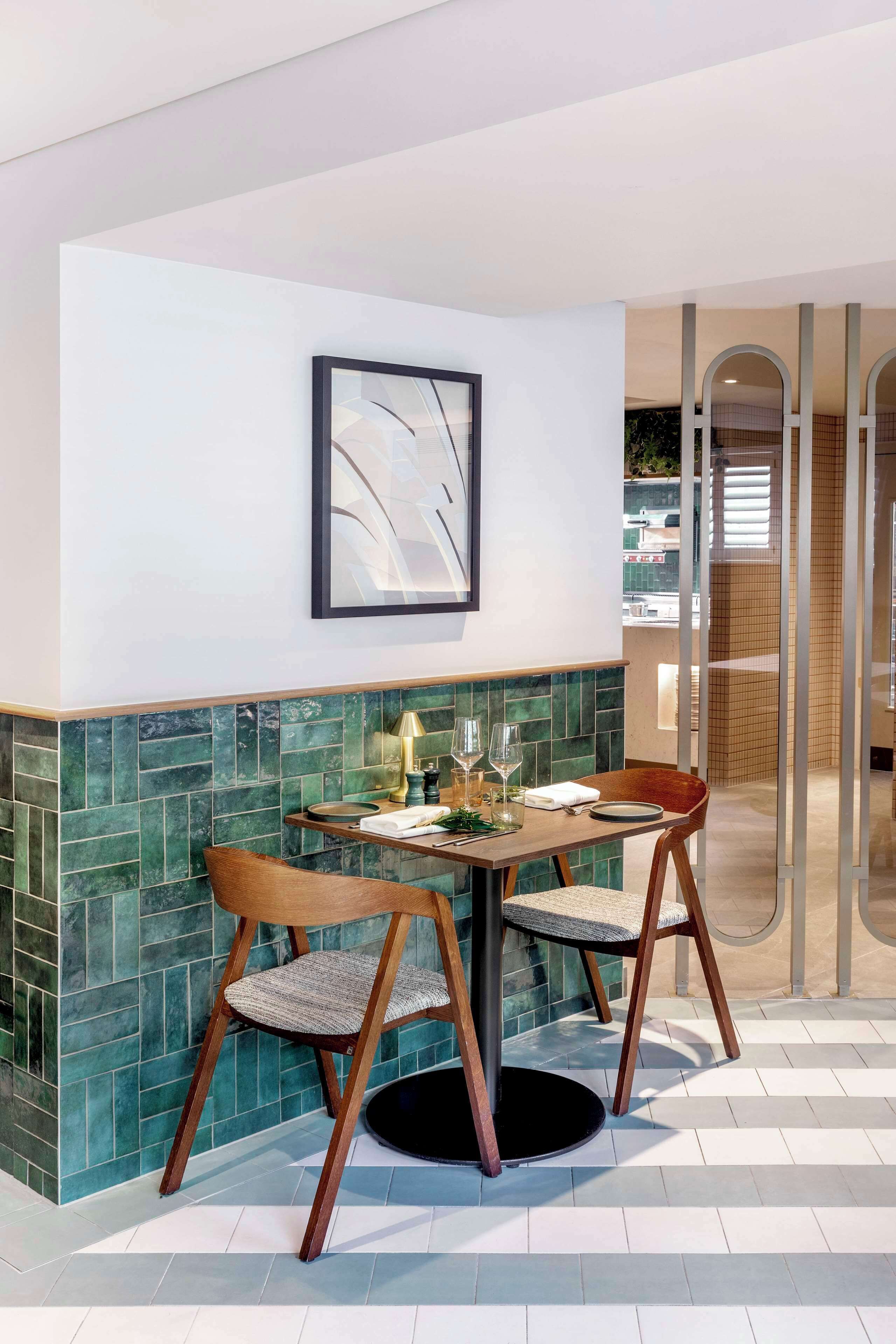 A dining area with a round table, two chairs, a framed artwork on the wall, and a green-tiled backsplash.