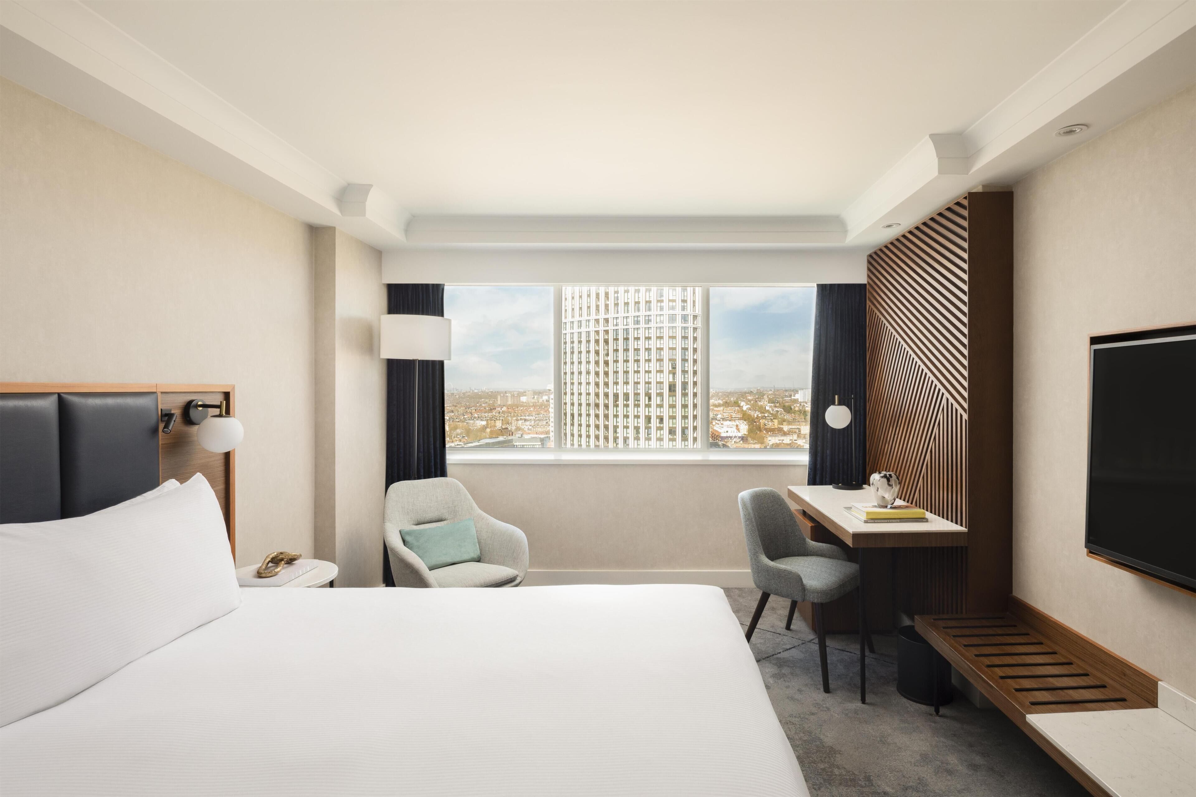 A hotel room with a large bed, a desk, two chairs, a TV, and a window with a city view.