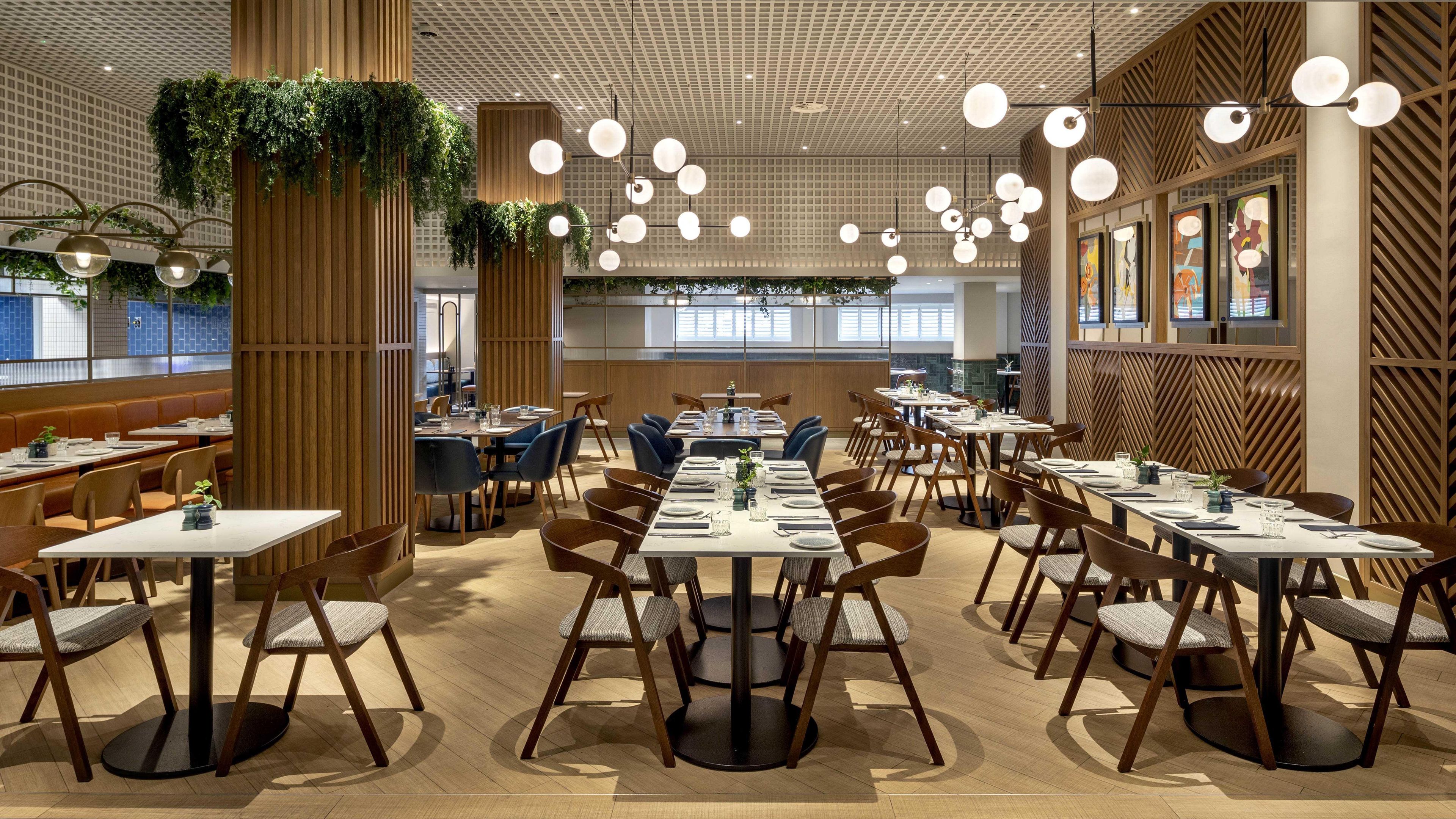 Ein modernes Restaurant mit mehreren Tischen und Stühlen, Holzelementen und hängenden Pflanzen.