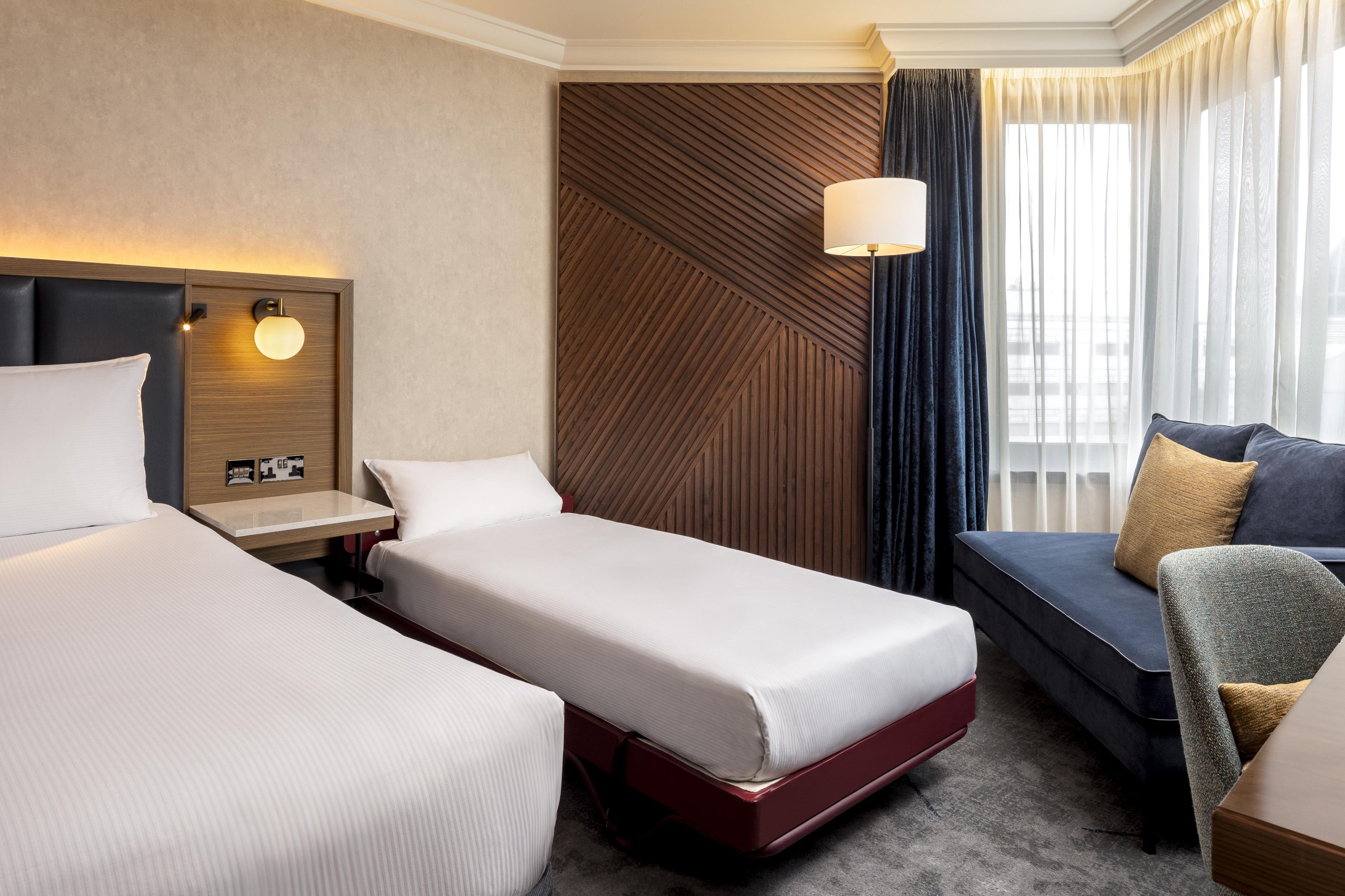A hotel room with two single beds, a sofa, a desk with a lamp, and a view of the city through large windows.