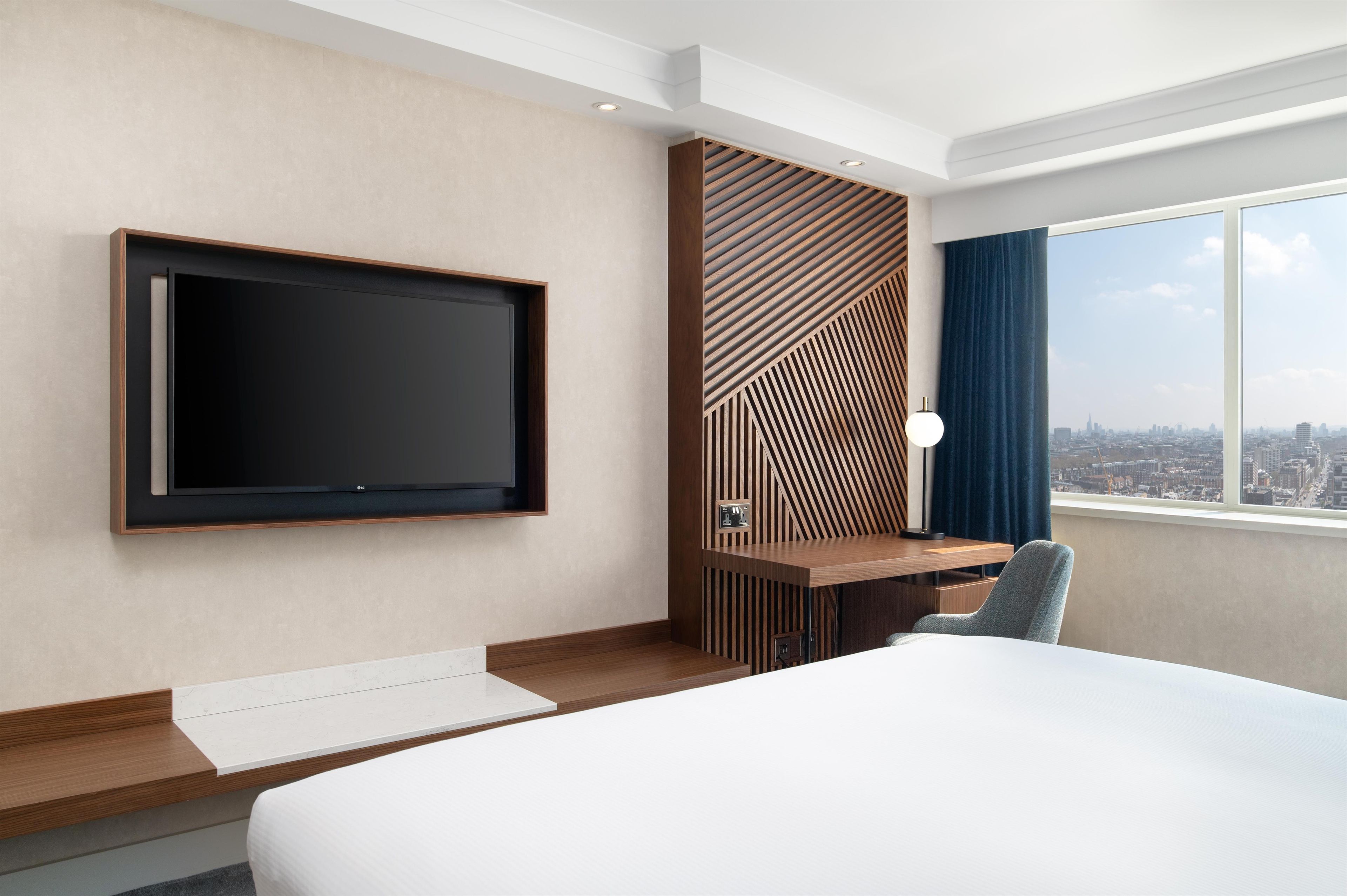 Una habitación de hotel con una cama, un televisor montado en la pared, un escritorio con una silla y vistas a la ciudad a través de la ventana.