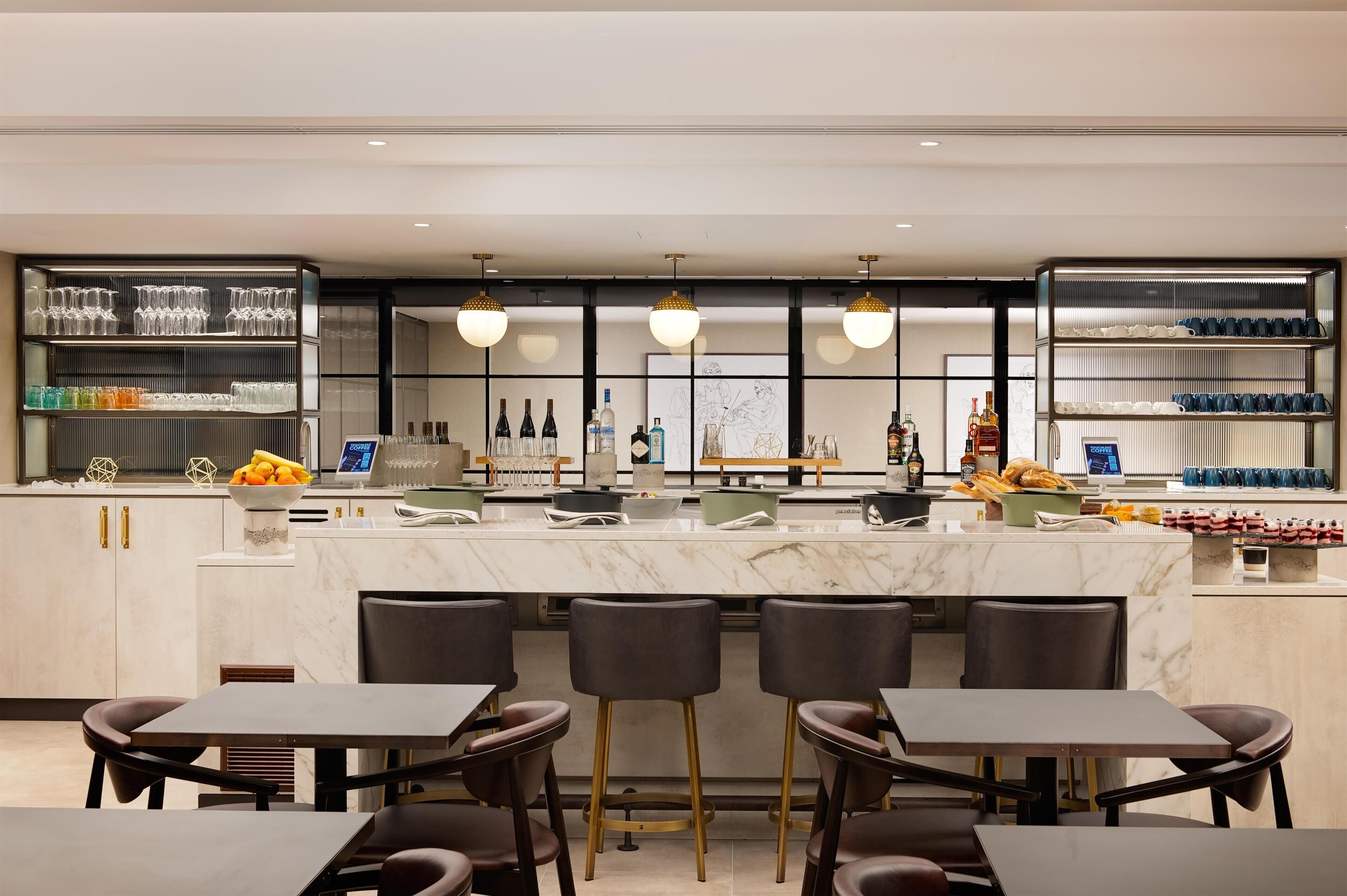 A modern bar area with marble countertops, bar stools, and shelves stocked with glasses and bottles.
