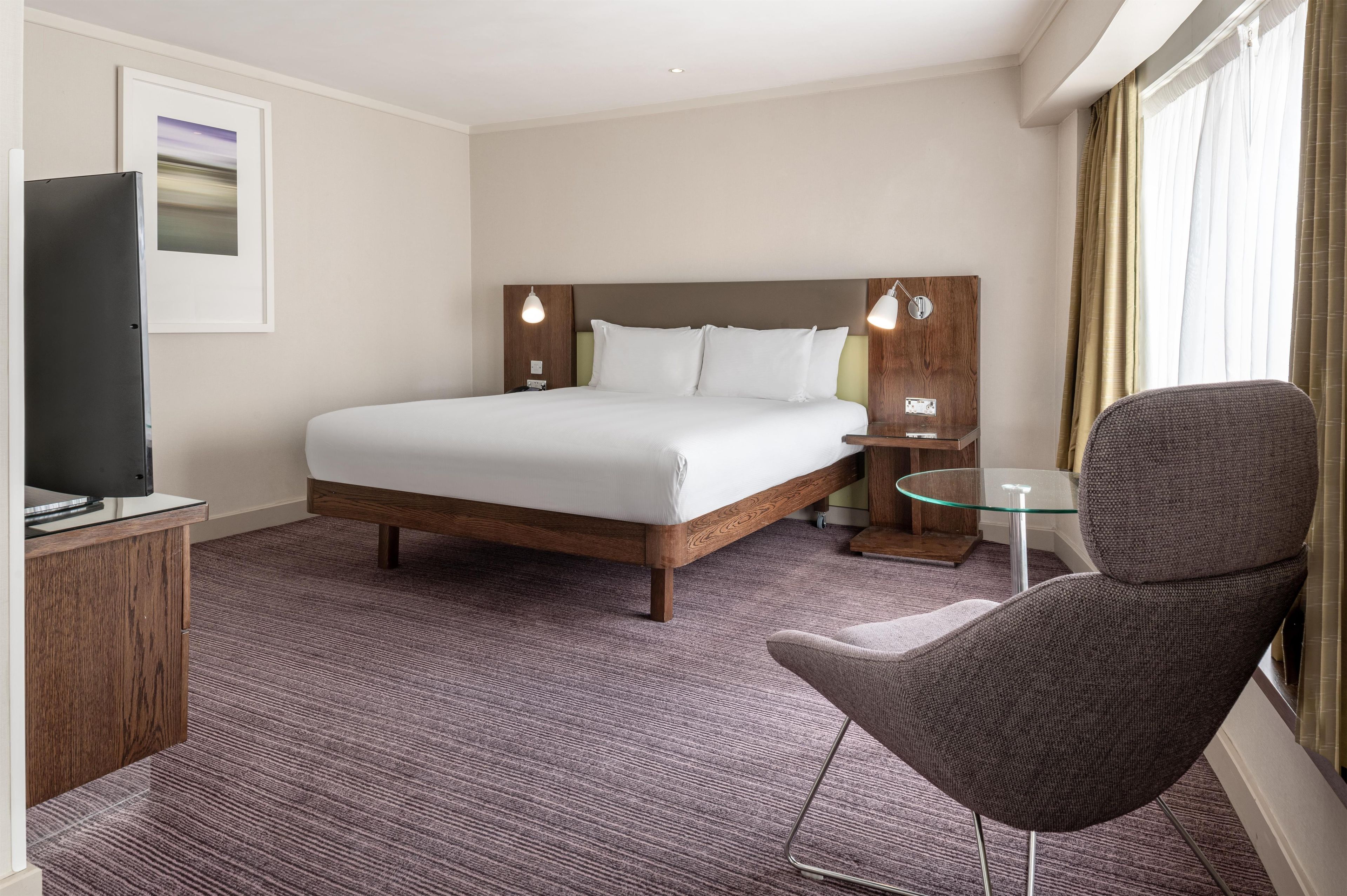 Ein Hotelzimmer mit Bett, Fernseher, Sessel, kleinem Tisch und einem Fenster mit Vorhängen.