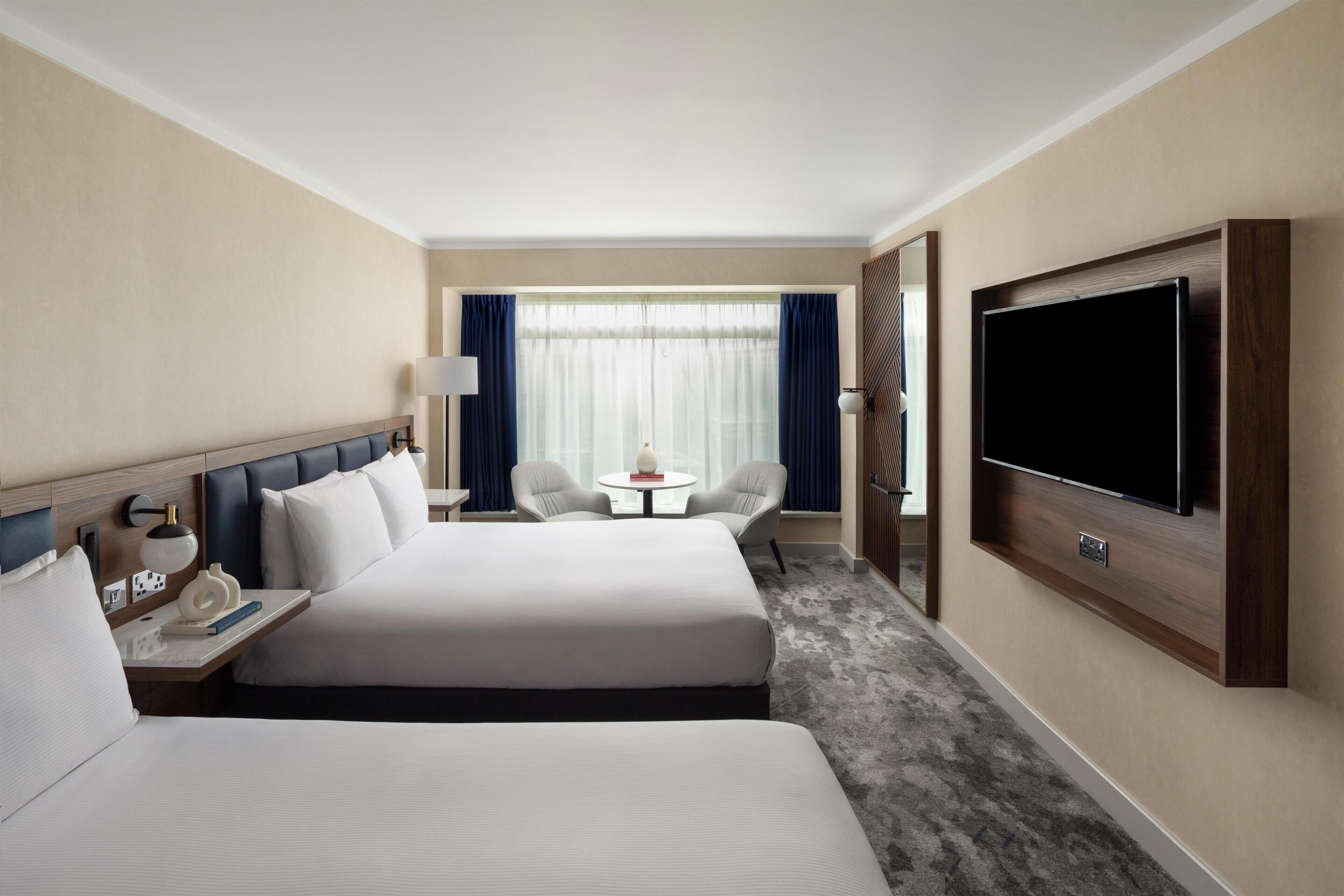A hotel room with two beds, a TV, a desk with a lamp, a small table, and a chair.