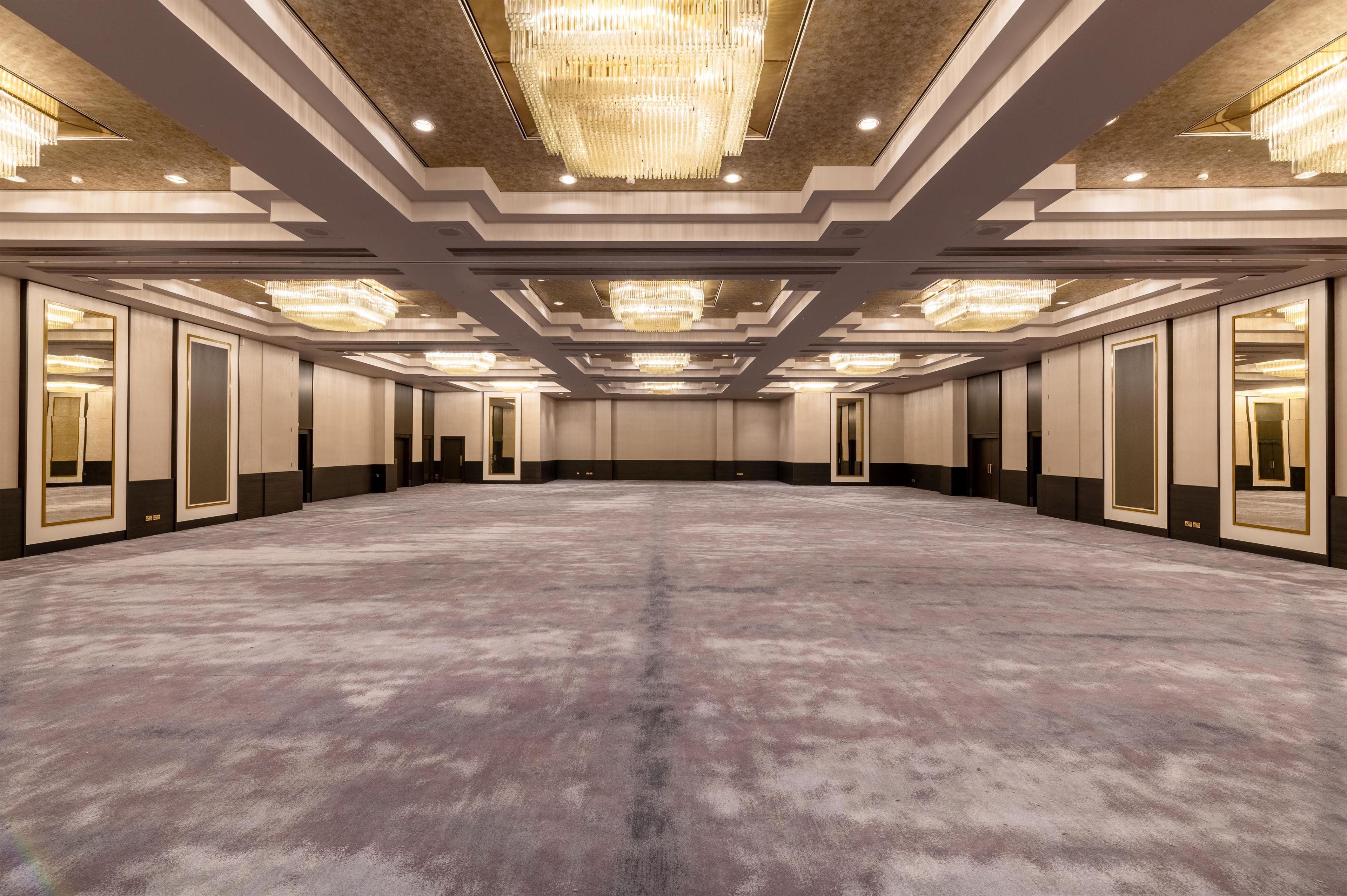 A spacious hotel conference room with a large carpeted floor, multiple doors, and a ceiling with chandeliers.
